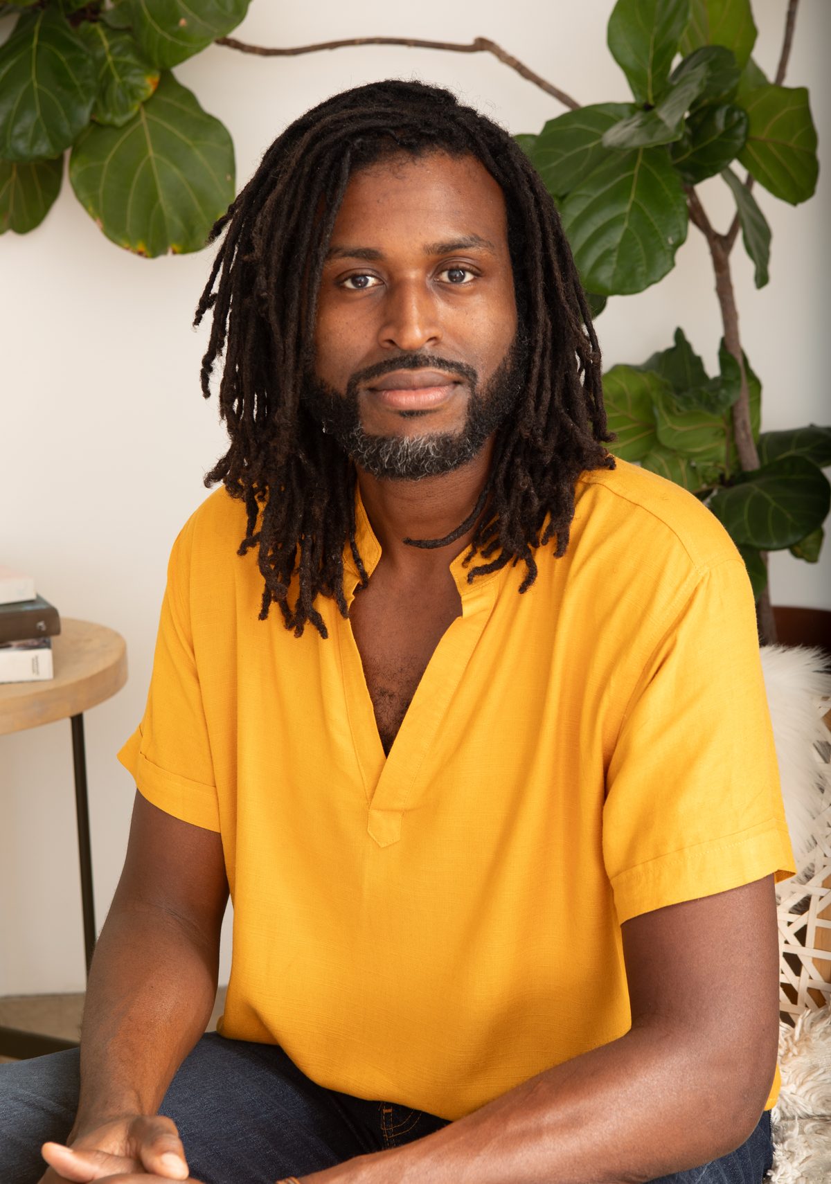 David Geathers seated in a bright room, locs down, in a golden-yellow shirt, a fiddle-leaf fig behind him.